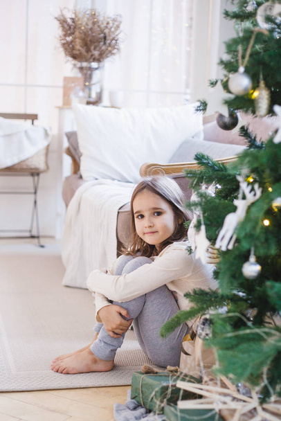 The girl is sitting near the sofa and decorated Christmas tree, the room is decorated for the new year and Christmas - Foto, Imagem