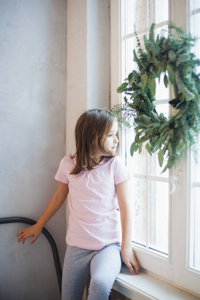 Menina de pé perto da janela esperando por Papai Noel, grinalda de Natal na janela, ano novo
 - Foto, Imagem