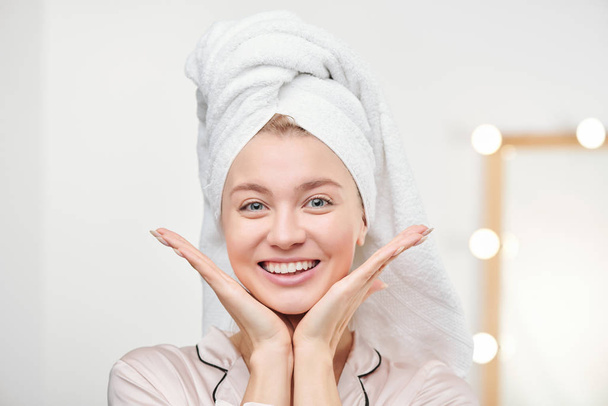 Chica bastante alegre con toalla en la cabeza disfrutando de su piel sana mientras mantiene las palmas abiertas por la cara después de la higiene matutina
 - Foto, Imagen