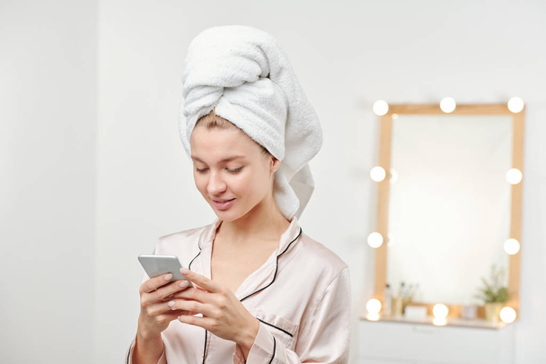 Młoda czysta kobieta z miękkim białym ręcznikiem na głowie przewijanie wiadomości w smartfonie po porannym prysznicu - Zdjęcie, obraz