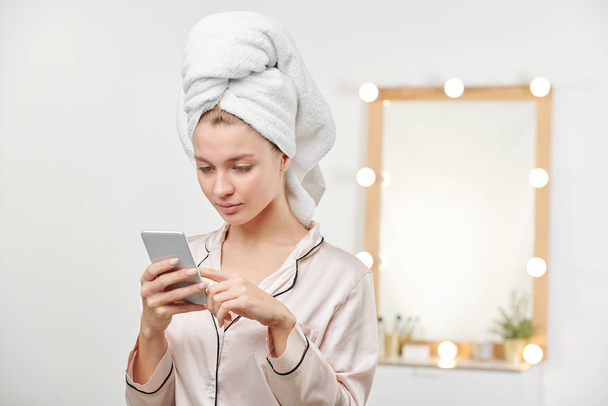Mujer joven y limpia en pijamas de seda desplazándose a través de mensajes en el teléfono inteligente después de la ducha de la mañana
 - Foto, imagen