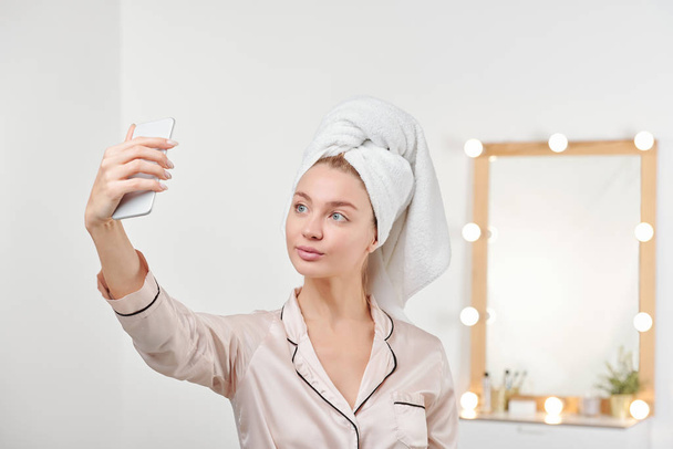 Świeża młoda kobieta w jedwabnej piżamie robiąca selfie po prysznicu stojąc na tle lustra z lampami - Zdjęcie, obraz