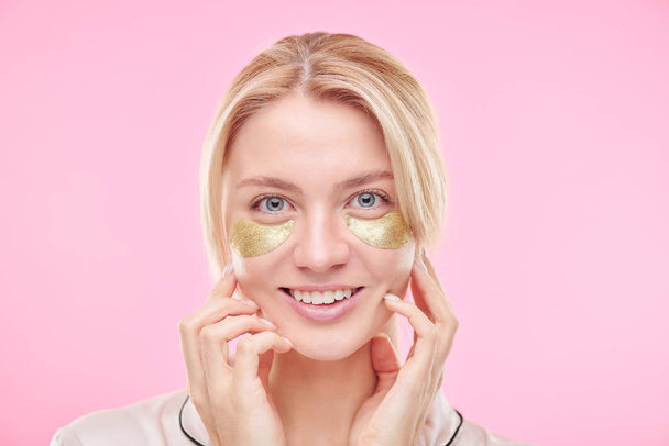 Smiling blond girl with golden revitalising under-eye patches touching her face while standing against pink background - Фото, изображение