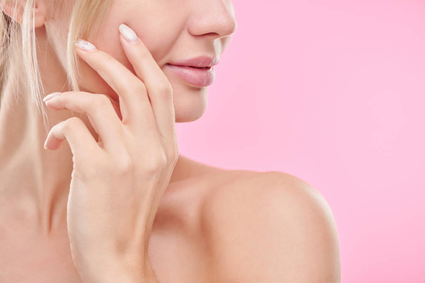 Mão de jovem mulher saudável tocando seu rosto enquanto cuida da pele contra fundo rosa em isolamento
 - Foto, Imagem