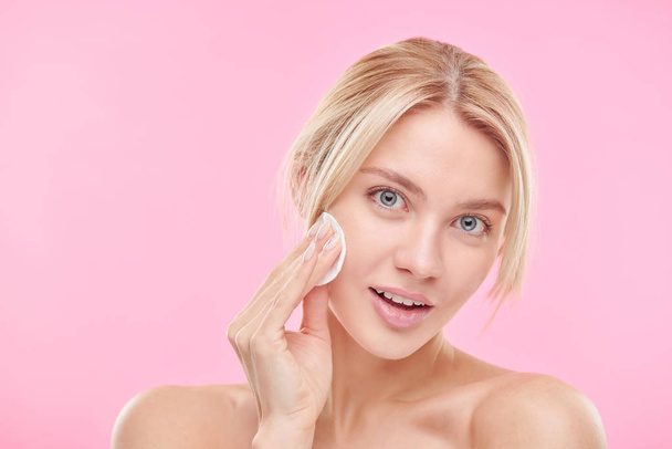 Menina loira bonita usando almofada de algodão durante a limpeza do rosto e remoção de maquiagem sobre fundo rosa
 - Foto, Imagem