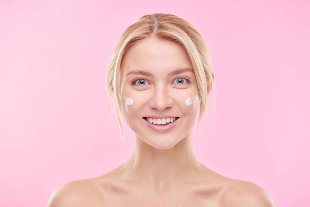 Happy blond girl with hydrating cream on her cheeks taking care of her face in front of camera in isolation - Foto, Imagen