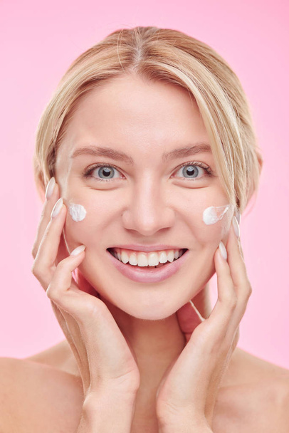 Young cheerful woman with toothy smile pampering cream on her face after cleaning it with toner - Foto, immagini