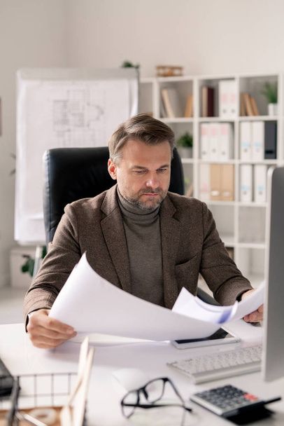 Poważny dojrzały architekt w stroju formalnym przeglądający papiery lub plany ze szkicami siedząc przy biurku w biurze - Zdjęcie, obraz