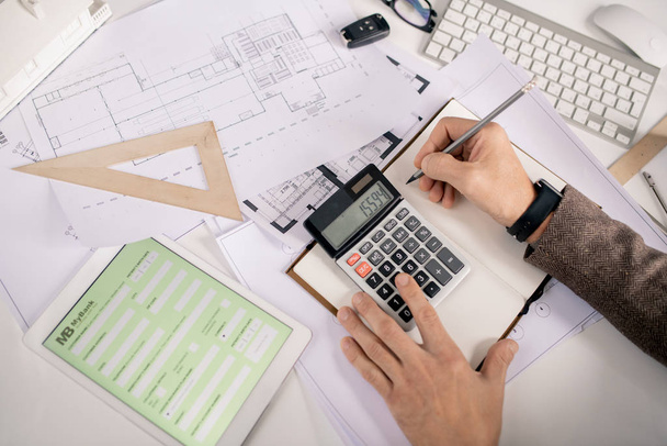 Вид рук инженера с ручкой и калькулятором, делающих заметки в ноутбуке во время работы за столом в офисе
 - Фото, изображение