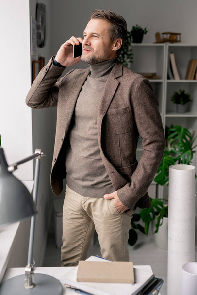 Уверенный зрелый архитектор со смартфоном, стоящим у окна офиса во время консультаций с коллегой или клиентом
 - Фото, изображение