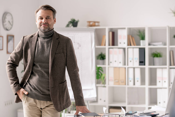 Confident mature architect in formalwear standing by desk in office on background of shelves with documents - Photo, Image