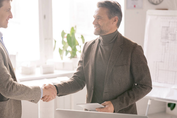 glücklicher reifer Ingenieur mit Touchpad begrüßt neuen Geschäftspartner per Handschlag nach Verhandlung im Amt - Foto, Bild
