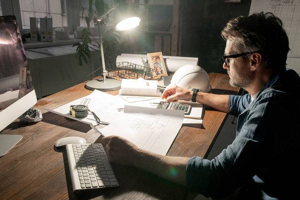 Współczesny inżynier lub przedsiębiorca siedzący przy stole i pracujący ze szkicami do późna w biurze - Zdjęcie, obraz