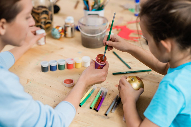 Творческая школьница с кистью берет краску или гуашь из маленькой пластиковой банки, расписывая на уроке золотой рождественский игрушечный мяч - Фото, изображение