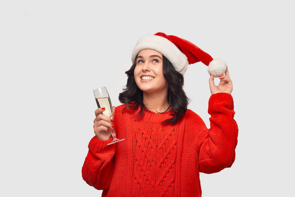 Beautiful brunette girl in a red sweater and Santa hat holding glass of champagne. - Photo, Image