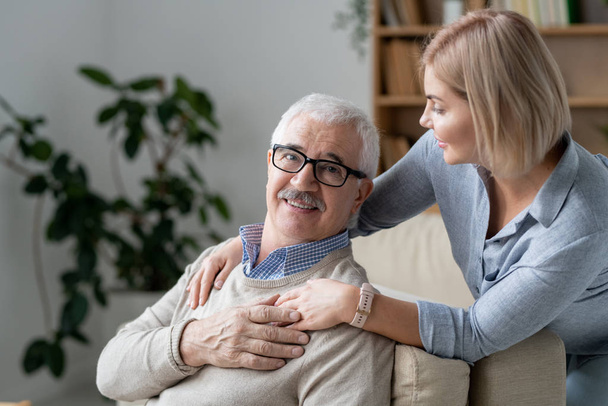 Успокаивающий пожилой человек на диване, держа за руку свою молодую блондинку дочь, стоящую рядом и обнимающую своего отца
 - Фото, изображение