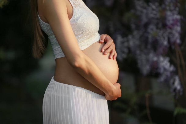Μια έγκυος γυναίκα σε προχωρημένη εγκυμοσύνη στον κήπο της Wisteria in bloom αγκαλιάζει την κοιλιά - Φωτογραφία, εικόνα