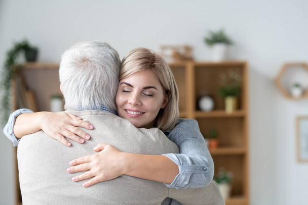 Молода блондинка ласкава жінка з зубною посмішкою і закритими очима, обіймаючи свого старшого батька в домашньому середовищі
 - Фото, зображення