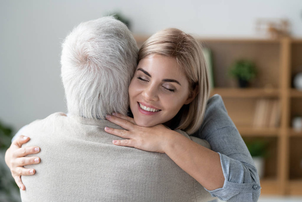 glückliche junge zärtliche Frau mit zahmem Lächeln umarmt ihren älteren Vater und drückt gleichzeitig Liebe und Fürsorge aus - Foto, Bild