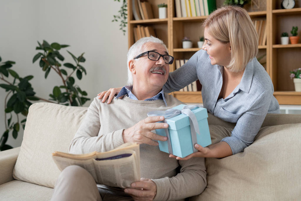 Ευτυχισμένη νεαρή περιστασιακή γυναίκα δίνοντας στον ηλικιωμένο πατέρα της συσκευασμένα γενέθλια ή χριστουγεννιάτικο δώρο σε giftbox, ενώ κοιτάζοντας τον με χαμόγελο - Φωτογραφία, εικόνα