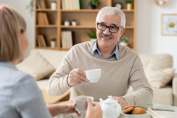 Happy retired man with cup of tea looking at his daughter during conversation by table in home environment - Foto, afbeelding