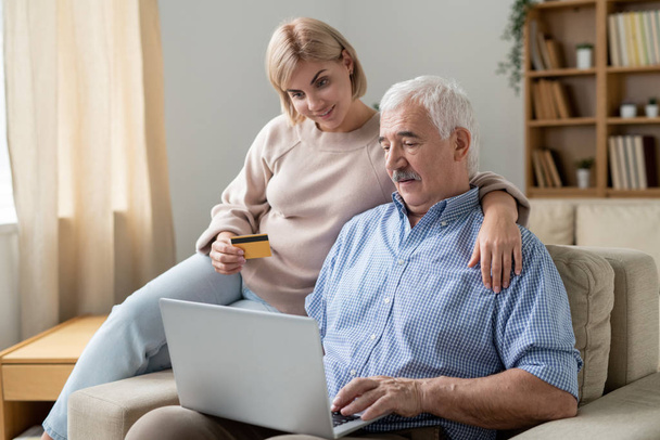 Молодая женщина с кредитной картой обнимает своего старшего отца с ноутбуком во время покупок онлайн на диване дома
 - Фото, изображение