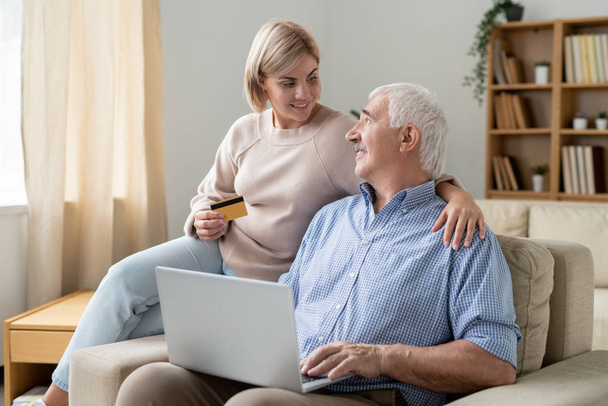 Ziemlich lässige Frau mit Kreditkarte schaut ihren pensionierten Vater mit Laptop an, während beide zu Hause online einkaufen - Foto, Bild