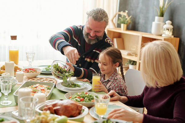 Happy grandpa giving salad to his granddaughter by festive table during Thanksgiving or Christmas family dinner at home - Photo, Image