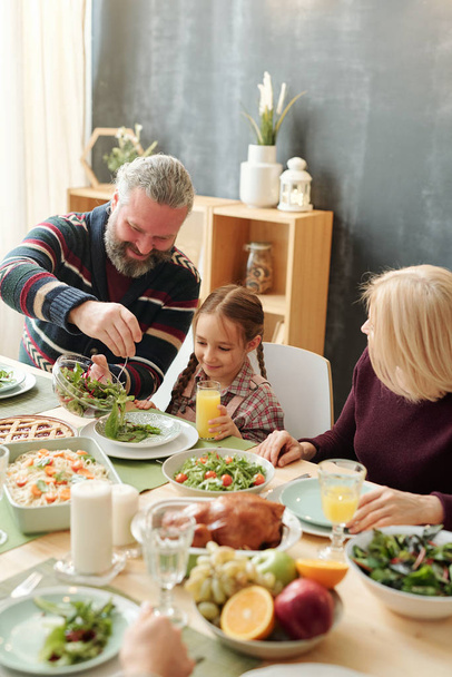 Happy mature man giving salad to his adorable granddaughter by festive table during family dinner on winter holiday - Photo, Image