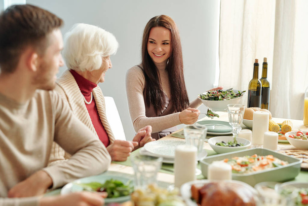 Smiling young woman putting salad on plate of grandma while looking at her by festive table during family dinner - 写真・画像