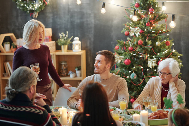 Members of big family looking at mature blonde woman with glass of wine making Christmas toast by festive dinner - Фото, изображение
