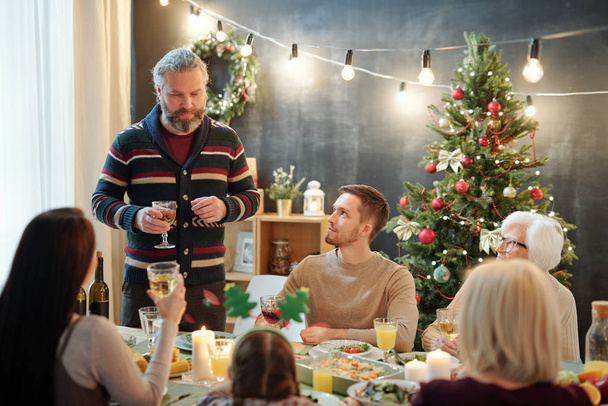 Bearded mature man with glass of wine toasting by served table while standing in front of his family during festive dinner at home - Фото, изображение