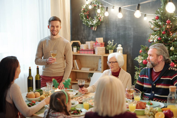 Χαρούμενος νεαρός άνδρας με ένα ποτήρι κρασί να ψήνει δίπλα στο τραπέζι μπροστά στην οικογένειά του κατά τη διάρκεια του Χριστουγεννιάτικου δείπνου στο σπίτι - Φωτογραφία, εικόνα