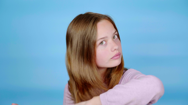 Teen girl in a pink pullover is combing her hair with a comb and looking at the camera. Blue background with copy space. In front of the mirror. Teenager emotions. 4k footage - Filmmaterial, Video