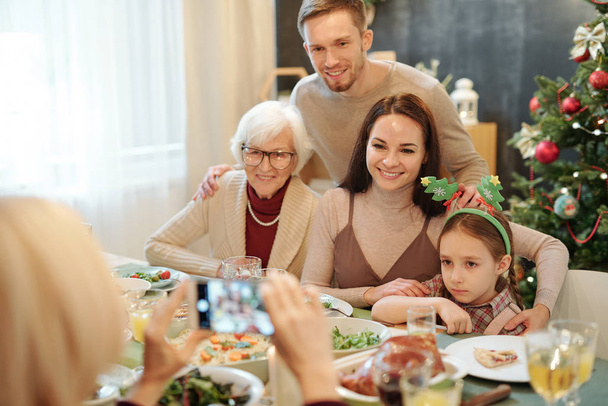 Famiglia affettuosa allegra seduta vicino al tavolo festivo servito e guardando la fotocamera dello smartphone nelle mani di una donna matura
 - Foto, immagini