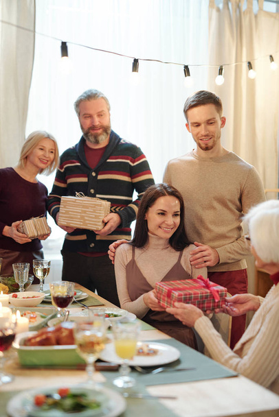 Jeune couple affectueux donnant cadeau de Noël dans une boîte cadeau à mamie assis près de la table de fête servie après le dîner
 - Photo, image