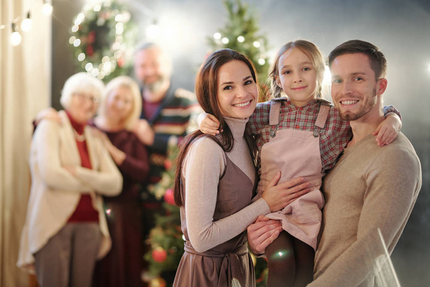 Happy young family of three standing in front of camera on background of mature adults by Christmas tree - 写真・画像