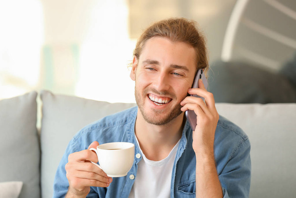 Handsome man with cup of coffee talking by mobile phone at home - Фото, зображення
