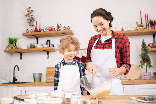 Happy young mother whisking raw eggs in bowl with her little son standing near by while helping his mom - Photo, Image