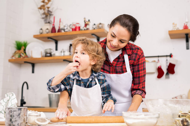 Roztomilý malý chlapec ochutnávka domácí těsto a zároveň pomáhá mamince, aby se válí na stole před pečivem nebo sušenky - Fotografie, Obrázek