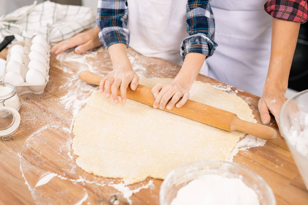 Руки ребенка с деревянной булавкой катятся свежее тесто на стол, помогая маме с приготовлением теста
 - Фото, изображение