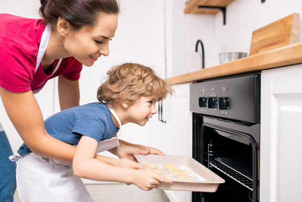 Giovane femmina aiutando il suo piccolo figlio a mettere vassoio con biscotti crudi in forno aperto mentre entrambi si piegano in avanti
 - Foto, immagini
