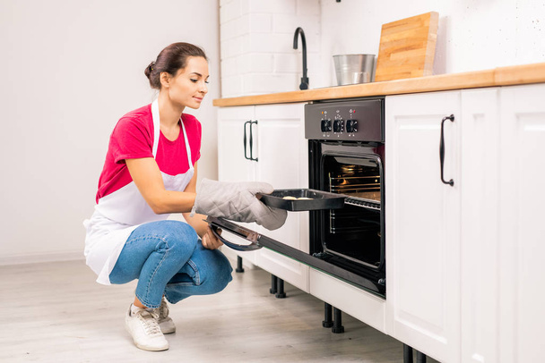 Giovane casalinga in grembiule accovacciata mentre mette vassoio con biscotti crudi nel forno elettrico mentre in cucina
 - Foto, immagini