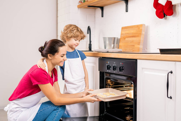 Στοργικό μικρό αγόρι στέκεται δίπλα στην ευτυχισμένη μητέρα του βάζοντας δίσκο με μπισκότα στο φούρνο, ενώ το μαγείρεμα μαζί - Φωτογραφία, εικόνα