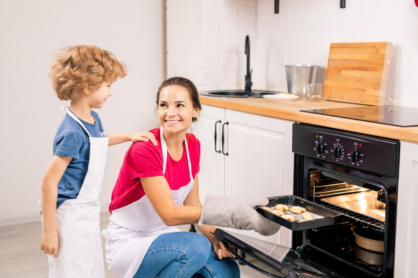 Cute boy in apron talking to his mom putting tray with raw cookies into oven while cooking together in the kitchen - Photo, Image