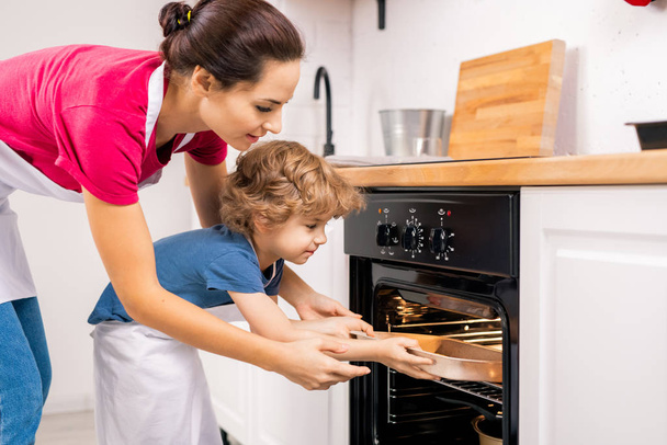 Kleiner Junge stellt Tablett in offenen Ofen und seine Mutter hilft ihm, nachdem sie am Wochenende selbst gebackene Plätzchen gebacken hat - Foto, Bild