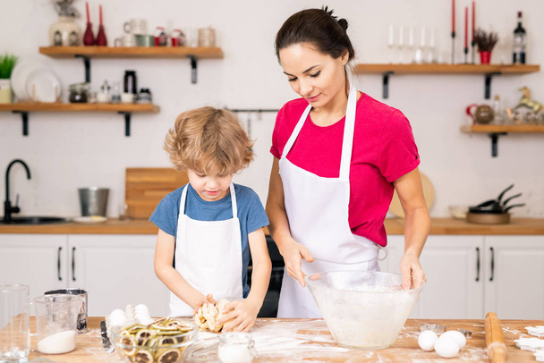 Uroczy chłopiec ugniatanie samodzielnie robione ciasto na ciasteczka, pomagając swojej mamie z gotowania przy stole w kuchni - Zdjęcie, obraz