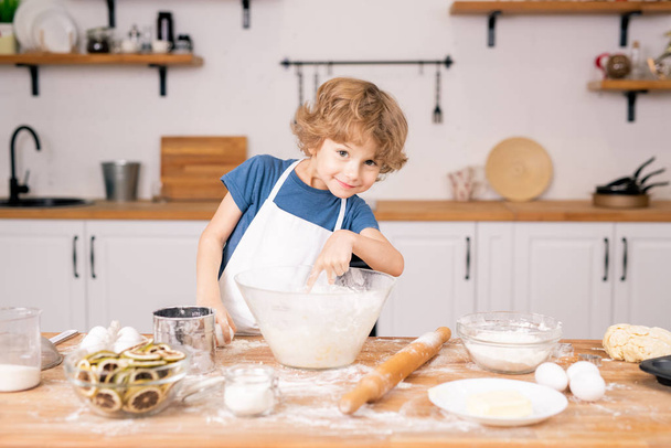 Słodki i zabawny mały chłopiec wskazując do miski z mąki, podczas gdy będzie łamać jajko, aby ciasto na ciasto - Zdjęcie, obraz
