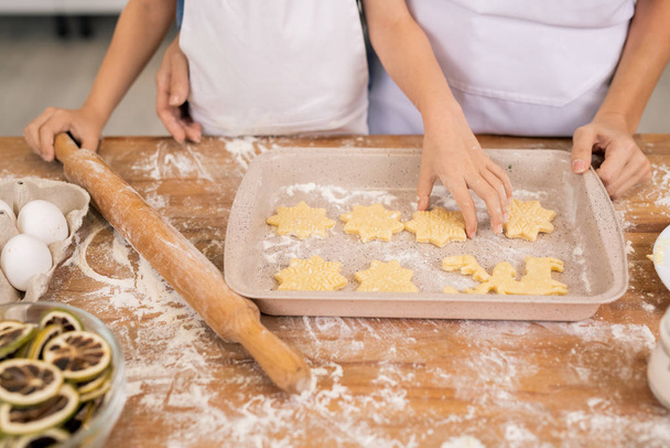 Handen van kleine jongen met roll-pin zetten rauw koekje in lade terwijl het helpen van moeder met het maken van koekjes - Foto, afbeelding