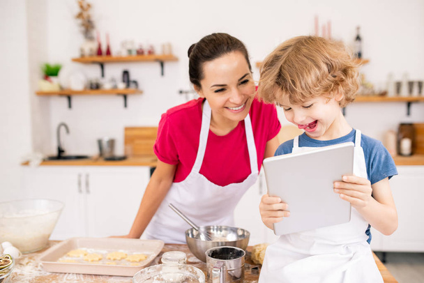 Очаровательный радостный ребенок с тачпад показывая своей маме видео рецепт чего-то действительно вкусный, выбирая, что готовить
 - Фото, изображение